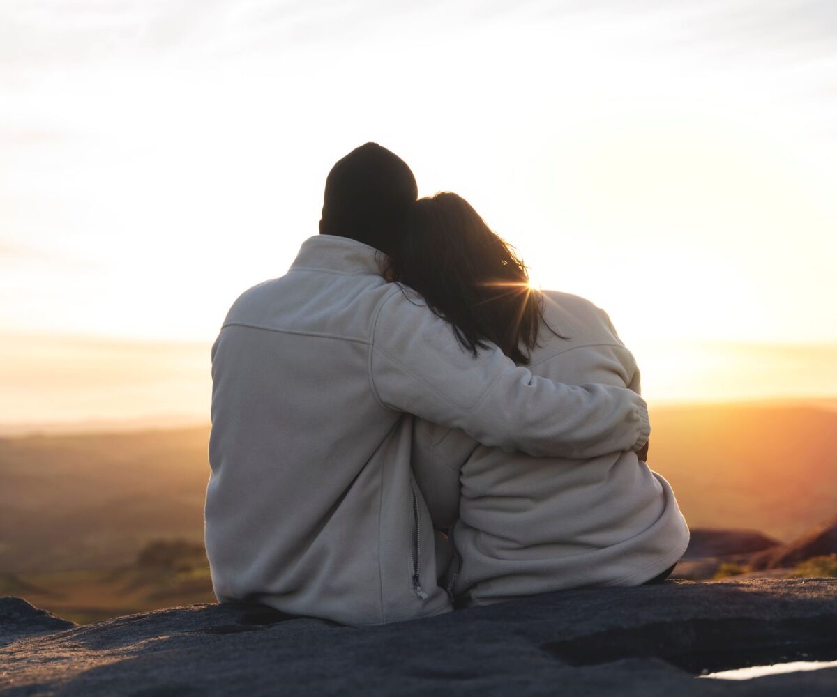 Back view of the happy couple in love sitting on top of a mountain enjoying a sunset landscape view