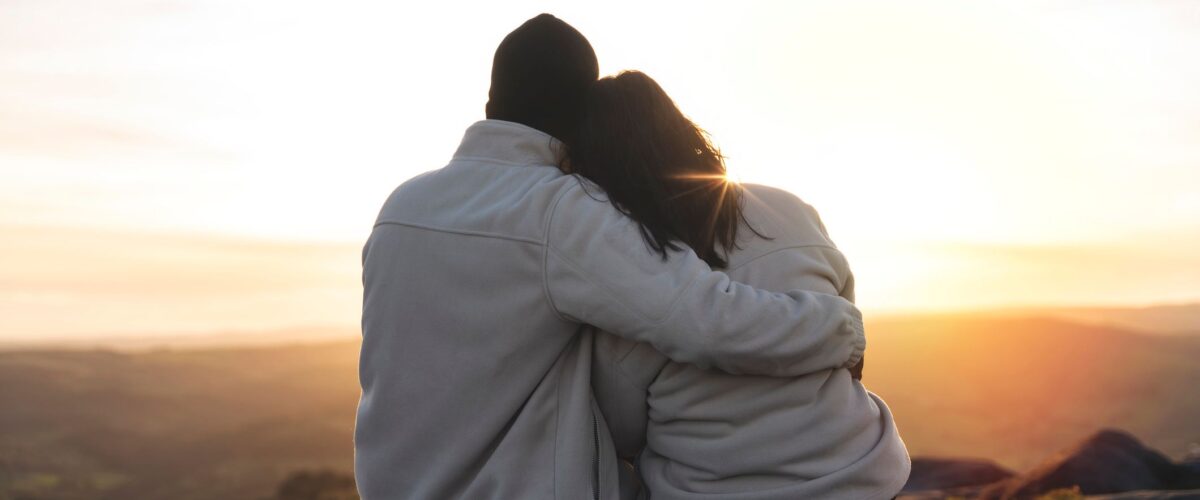 Back view of the happy couple in love sitting on top of a mountain enjoying a sunset landscape view